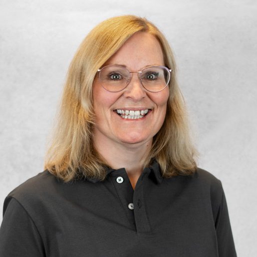 Lächelnde Frau mit blonden Haaren, Brille und dunklem Hemd vor grauem Hintergrund.