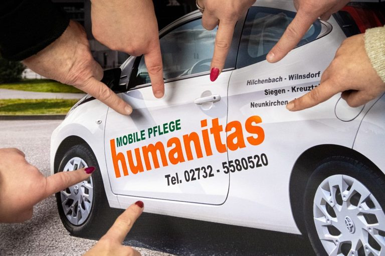 Zusammen versammelte Hände zeigen auf die Seitenansicht eines weißen Autos mit dem Schriftzug "humanitas".