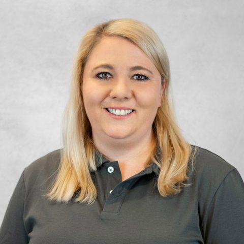 Blonde Frau mit lässigem Polohemd und freundlichem Lächeln vor neutralem Hintergrund.