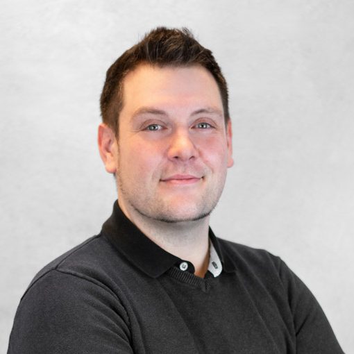 Mann in schwarzem Pullover, lächelnd mit verschränkten Armen vor grauem Hintergrund.