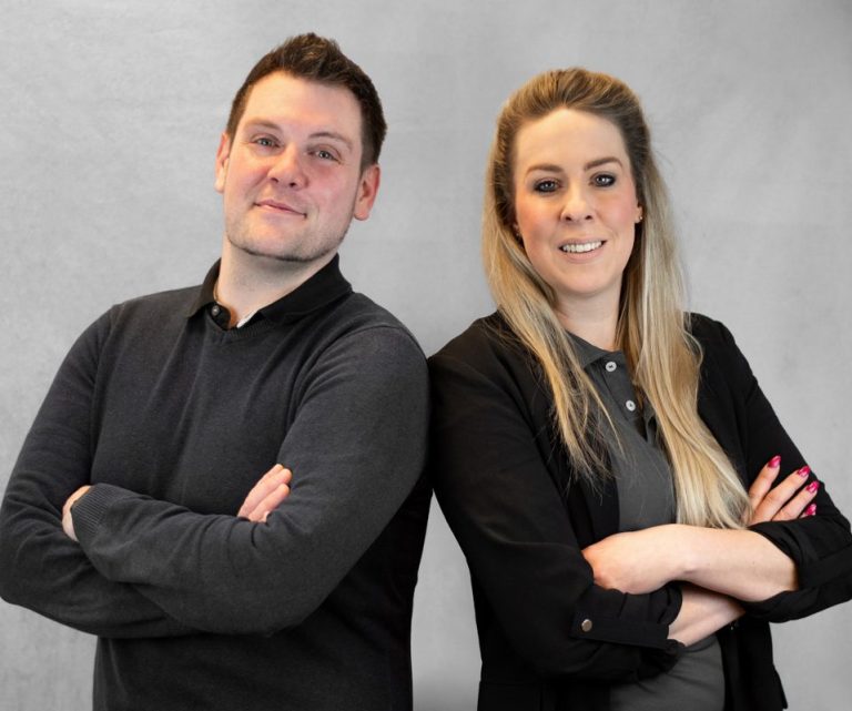 Die zwei Geschäftsführer der Mobile Pflege Humanitas GbR, Katrin und Sebastian, stehen mit verschränkten Armen vor einem grauen Hintergrund.